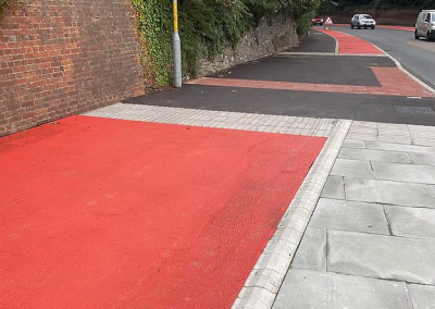Street view showing a cycle scheme with bright red surfacing laid along the carriageway and crossing points, running past historic brick walls and mature trees near St Augustine’s Abbey. The modern red tarmac contrasts sharply with the surrounding historic setting and materials.