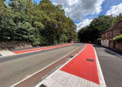 Street view showing a cycle scheme with bright red surfacing laid along the carriageway and crossing points, running past historic brick walls and mature trees near St Augustine’s Abbey. The modern red tarmac contrasts sharply with the surrounding historic setting and materials.