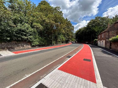 Street view showing a cycle scheme with bright red surfacing laid along the carriageway and crossing points, running past historic brick walls and mature trees near St Augustine’s Abbey. The modern red tarmac contrasts sharply with the surrounding historic setting and materials.